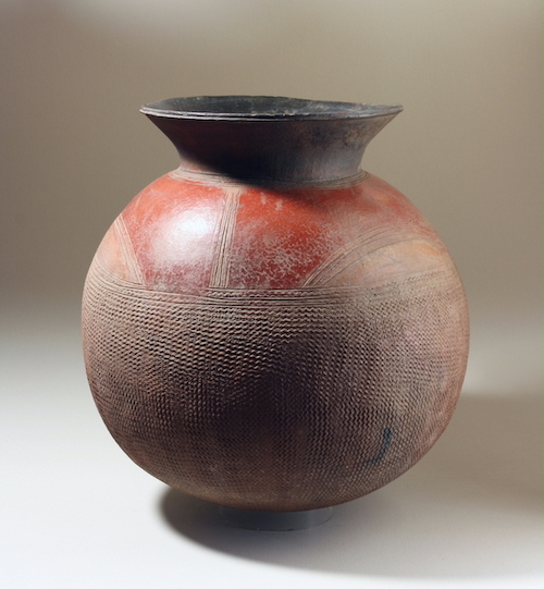 A vessel with a bulbous body and a tin slightly flaring neck. The body of the vessel is decorated with line designs. The vessel is mostly tan and red.