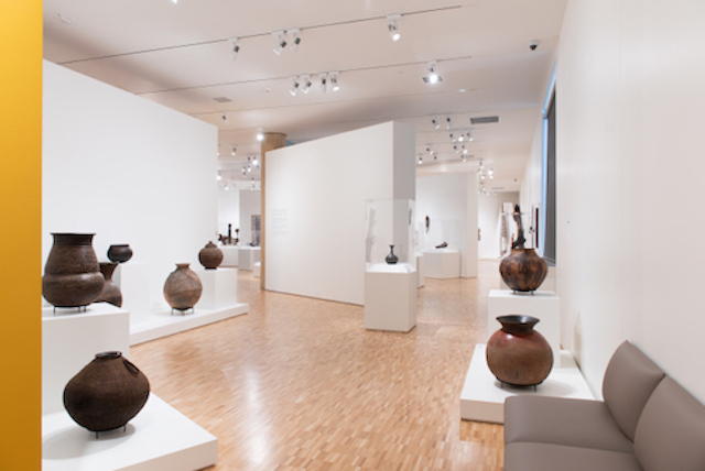 An installation view of a gallery, depicting an arrangement of brown ceramic pots in varying shapes and sizes.