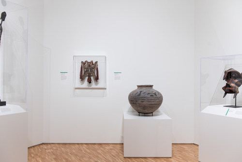 An view of a gallery installation, showing several art objects in glass cases and a large grayish ceramic pot.