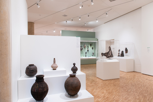 A view of a gallery installation. In the foreground is a display of five ceramic pots in various sizes and shapes.