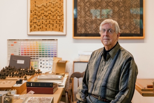 A white-haired, light-skinned man in a long-sleeved shirt is shown in a room that has art objects on tables and on the walls behind him.