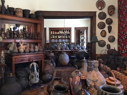 A home living space is shown. It is full of ceramic pottery in various sizes and shades of brown, which are on the floor, fireplace, and tables throughout the space.