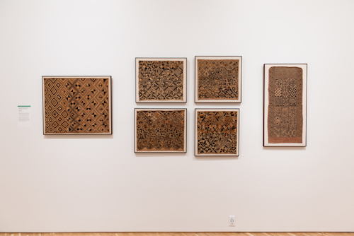 A view of six textiles in various shades of brown. All of the textiles are framed and installed on the wall of a gallery.