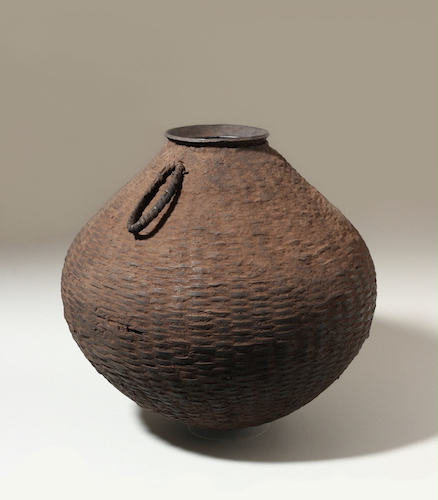 A rounded brown ceramic vessel with a narrow mouth, one loop-like handle, and a weave-like texture.