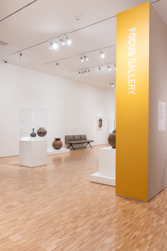 An view of a gallery, showing a yellow wall with the words 'Focus Gallery' in white and ceramics in various sizes on display.