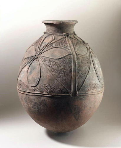 A tall, oval-like ceramic vessel with a narrow, flared mouth on top. It has raised shapes and textures around the body, including a four-leaf flower-like shape.