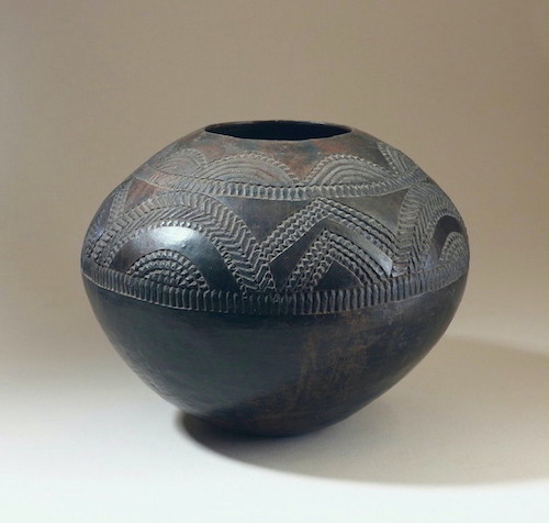 An ovoid vessel with a wide waist and a lipless rim. It is decorated with inscribed bands and patterns of semi-circular designs from the waist to the rim. The vessel has thin walls with a smooth glossy surface between cut designs.