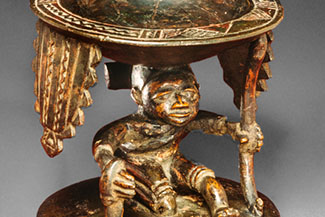 A divination cup with a seated male figure holding up the shallow bowl of the cup. The figure has large and prominant genitals and holds a staff in his left hand.