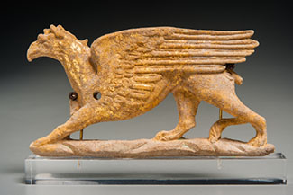 A griffin is carved in relief and faces left, standing on a ground line. The right paw and part of the tail are missing and a small hole is pierced through the shoulder. Details such as wing feathers, claws, and a heavy brow are carefully depicted.