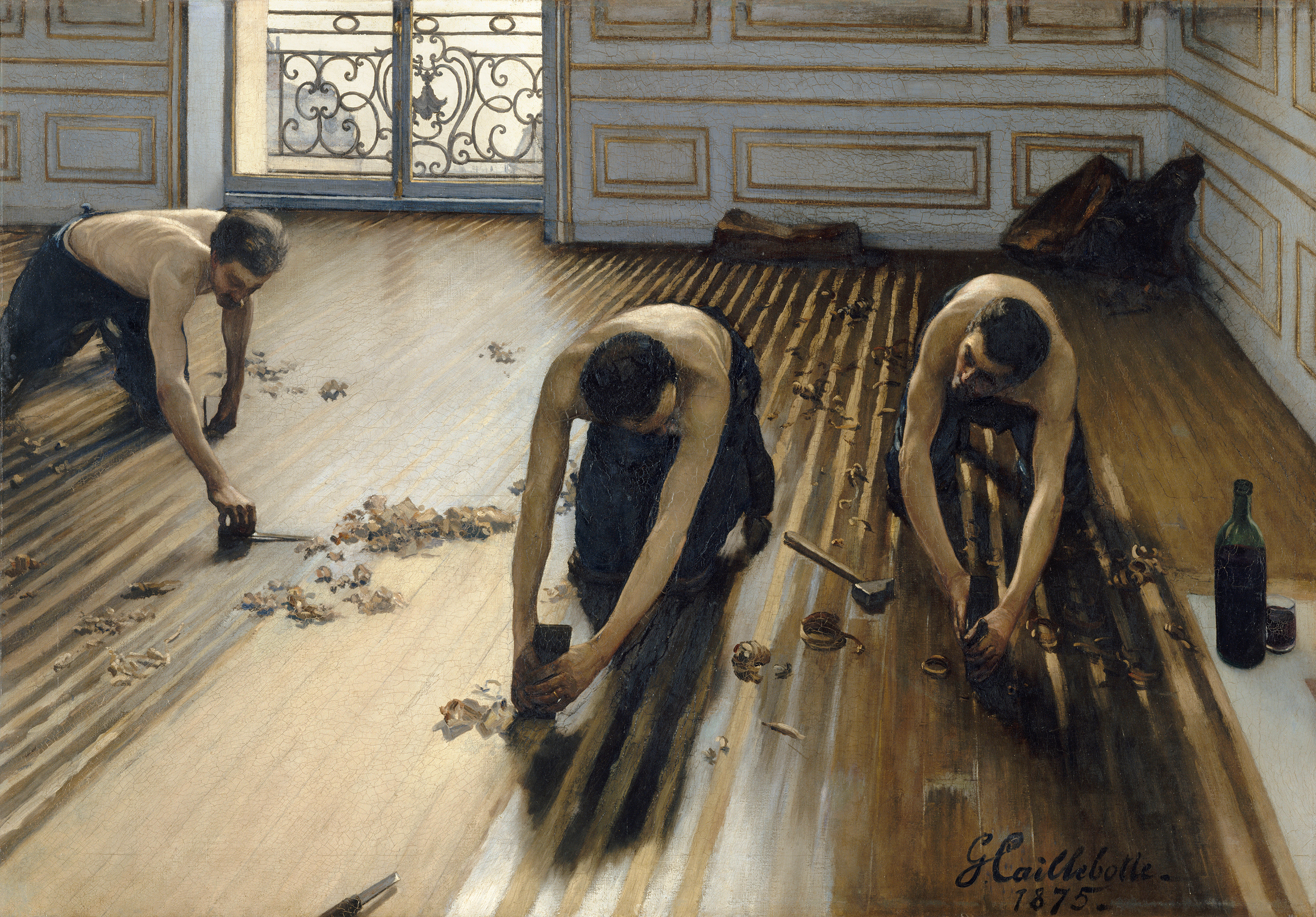 Three men kneel on a wood floor in a brightly lit room. They are scraping the wood of the floor, and curled wood shavings are around them.