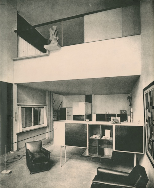 The furnished interior of a two-level modernist home is shown in a black-and-white photo. On the main level are two chairs, a tall three-sectioned sideboard with the center door opened down, and an abstract framed painting hanging on the right side. There is a lofted second level with a partially-glass half-height wall.