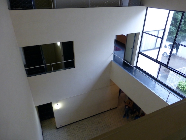 A building's interior atrium is shown from a slightly elevated angle, as if the photographer is standing on the second level of the building. The walls are white and feature geometric, rectangular windows.