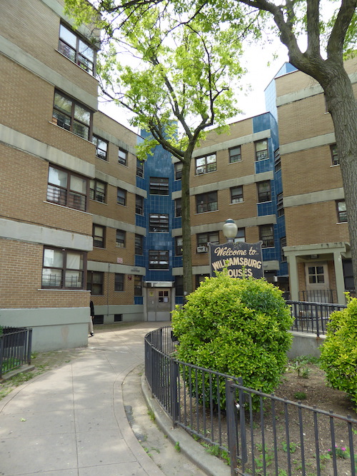 The front of an L-shaped apartment building is shown. It is primarily reddish-brown brick, with columns blue tile facing evenly spaced on the left, right, and corner of the L shape.In front of the building are fenced-in trees and small shrubs, along with a sign that reads 'Welcome to Williamsburg [H]ouses'