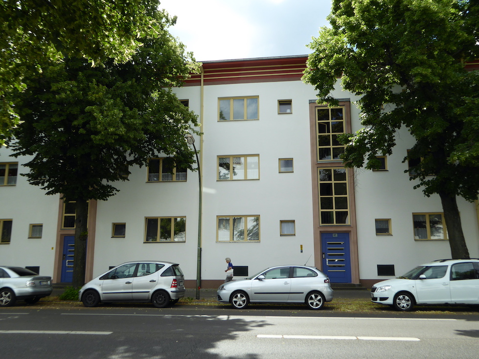 Three-story white buildings face a street. The buildings’ doors are blue and the windows are framed in yellow. Four cars are parked on the street in front of the buildings.