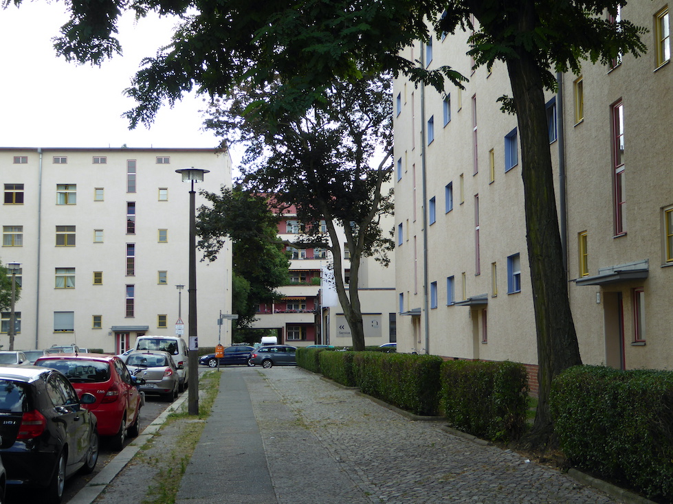 Four-story beige buildings are arranged around a sidewalk and street. The buildings’ facades contain red, yellow, and blue accents around the windows and doors. Two large trees and a row of hedges are planted to the right of the sidewalk.