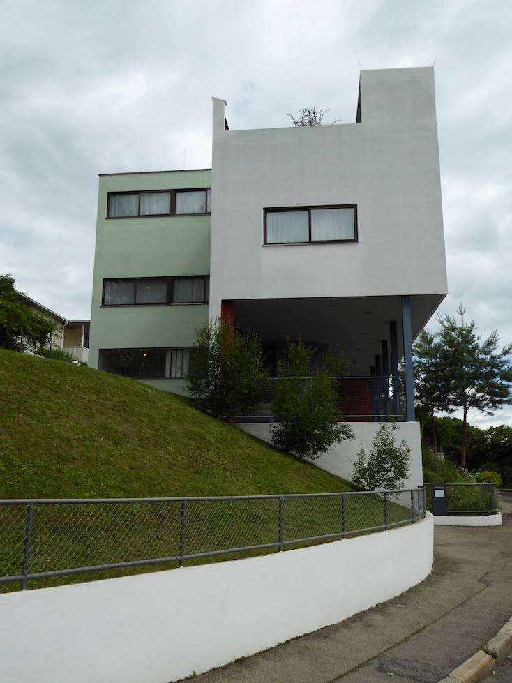 A white and pale green building stands behinds a grassy hill. The building is constructed of two cube-like structures. Four horizontal windows punctuate the façade. 