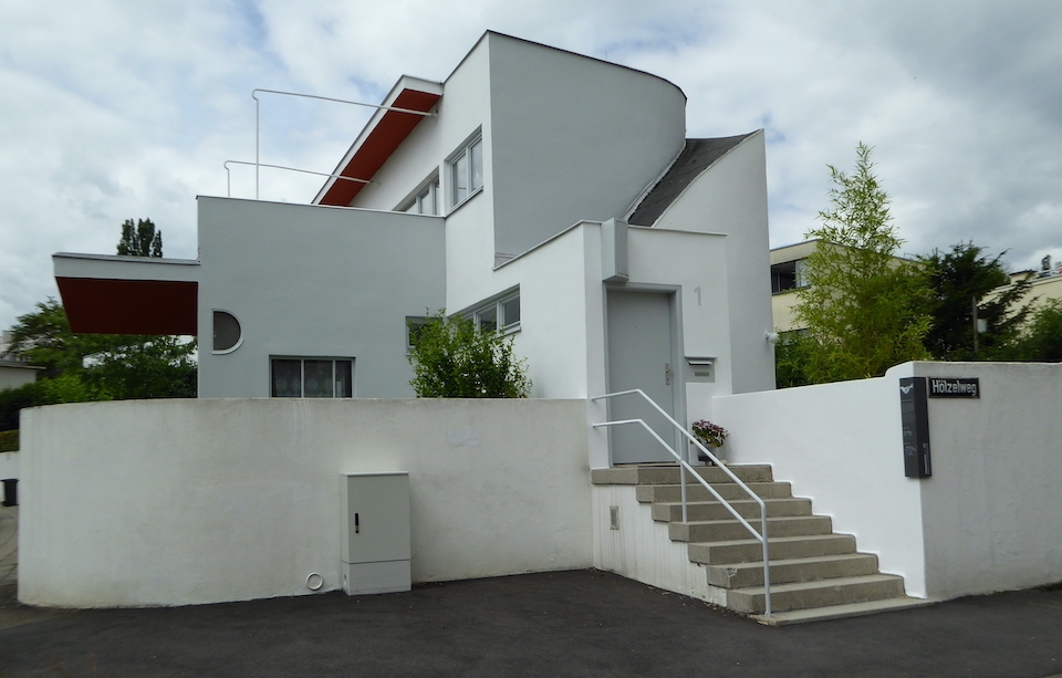 A geometric white house sits on a street corner. The house is constructed of interlocking cube-like and rounded shapes. A stairway with a white railing leads to the front door at the right.