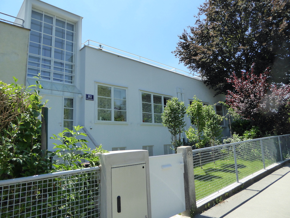 A long, white, two-story building stretches down a city block. At the left, a large glass window juts up above the roofline. A landscaped lawn with trees is separated from the sidewalk by a mesh fence.