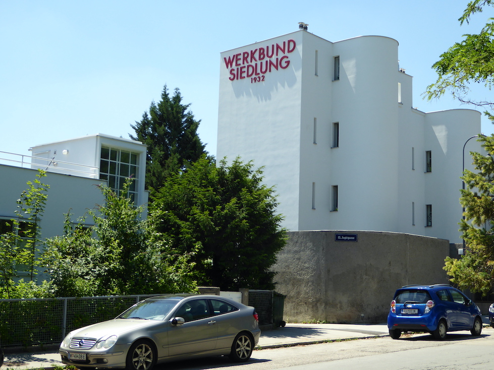 A large white building faces a street. Two rounded elements jut out from the flat façade. A shorter white building with a large window and a group of trees are situated to the left.