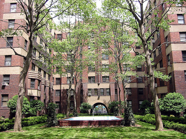 A U-shaped, red brick building is arranged around a grassy courtyard. Horizontal bands of tan stucco delineate each floor of the building. Four tall trees are planted in the courtyard.
