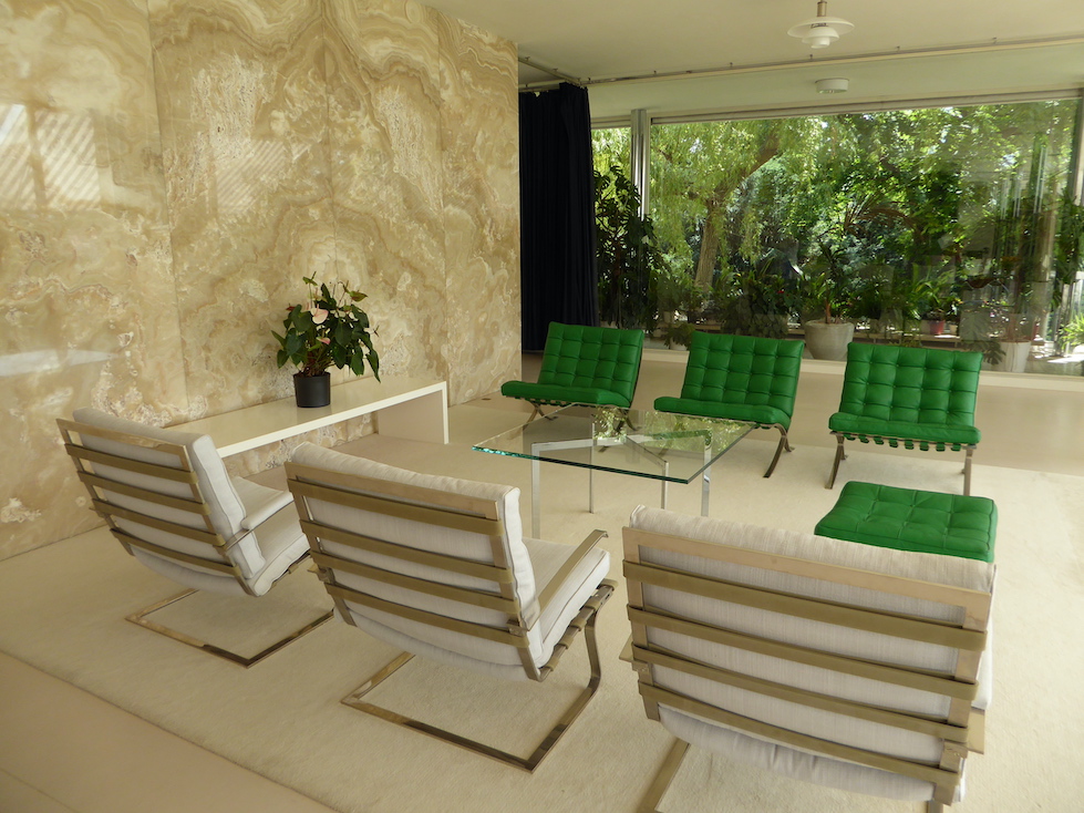 Six chairs arranged in two rows of three face each other in an interior face. Three chairs are upholstered in green and three in white. Dense green foliage is visible through a large plate glass window behind the chairs.
