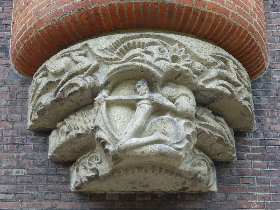 A white, rounded stone carving is attached to a gray and red brick façade. The carving depicts a simplified human form holding a bow and arrow. Decorative animal and flower forms are carved in relief above the archer.