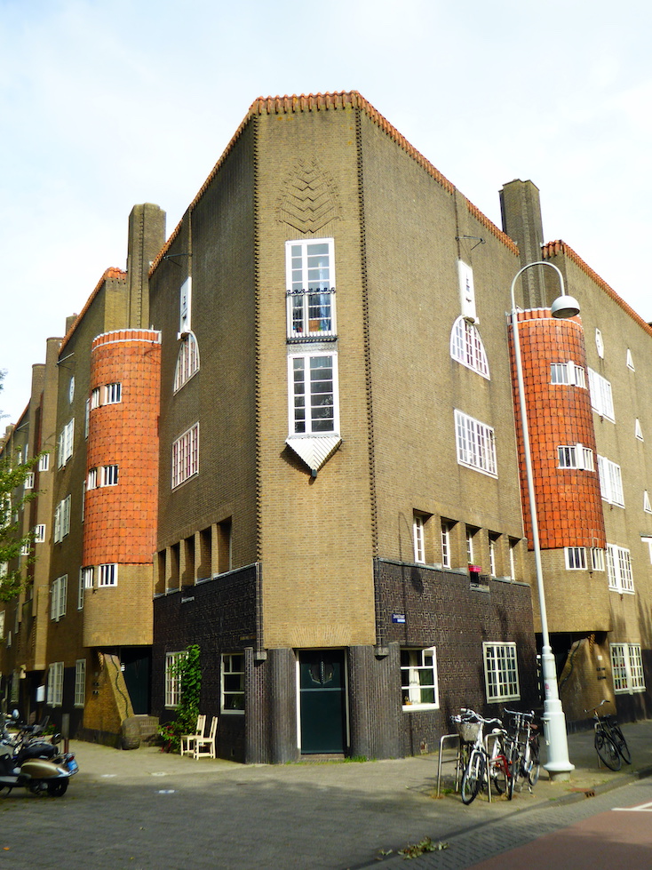 The corner of a large building faces a sidewalk. The building’s façade is decorated with tan and gray brick, red tile, and carvings. Two rounded architectural features frame the corner.