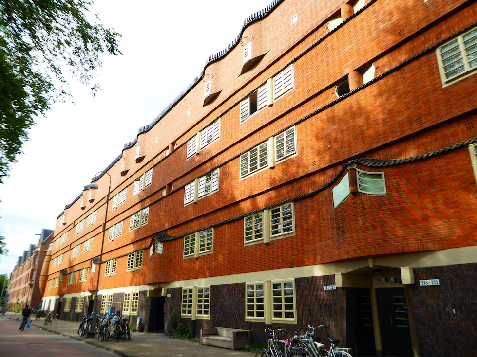 A large, five-story building stretches the length of a city block. The red and gray brick façade is punctuated with pointed and rounded protrusions.