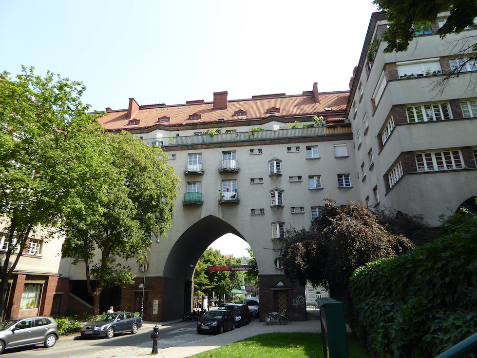 A six-story building is situated around a street. The façade is stucco and the roof is red tile. A pointed archway covers the street.