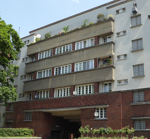 A building façade contains a central bank of windows. Decorative brickwork in a chevron pattern sits beneath the windows. Beneath this is a square archway.