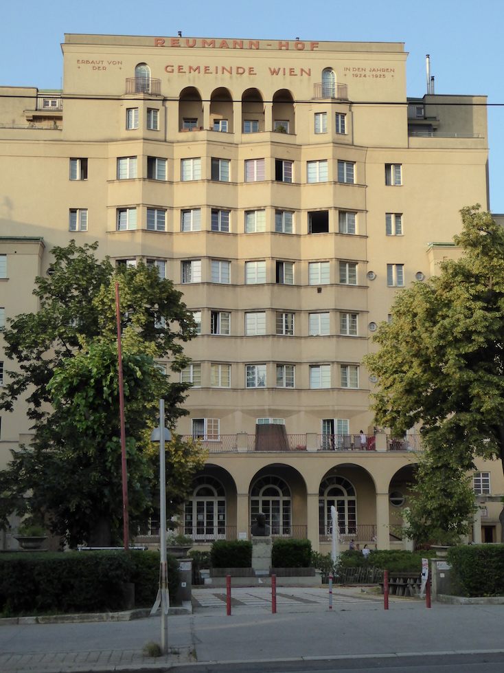 A tall yellow building faces a sidewalk. The façade features a central bank of windows arranged in an undulating formation. Trees are planted on both sides of the building’s entrance.