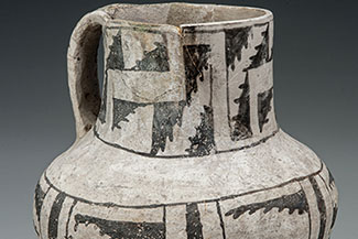 A clay pitcher with a narrow bottom, wide shoulders, and a wide, cylindrical neck. The vessel is mostly white in color, but geometric designs are painted in a grey/black slip on the lower portion of the vessel's body, and on the neck.
