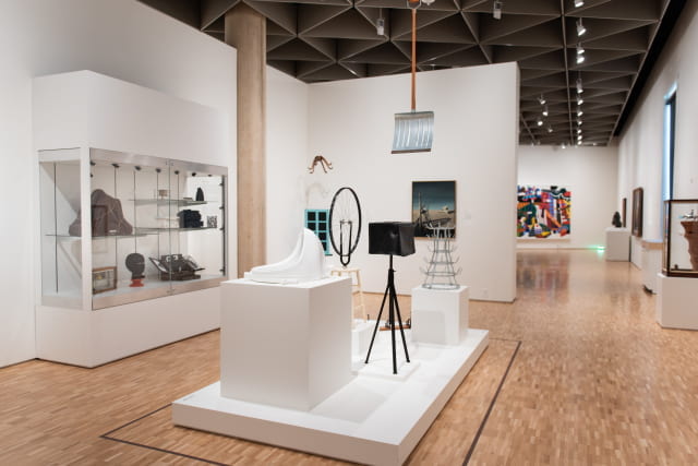 An installation view of Marcel Duchamp's Readymades, including a bicycle wheel mounted on a stool, a window with blue framing, and a urinal.