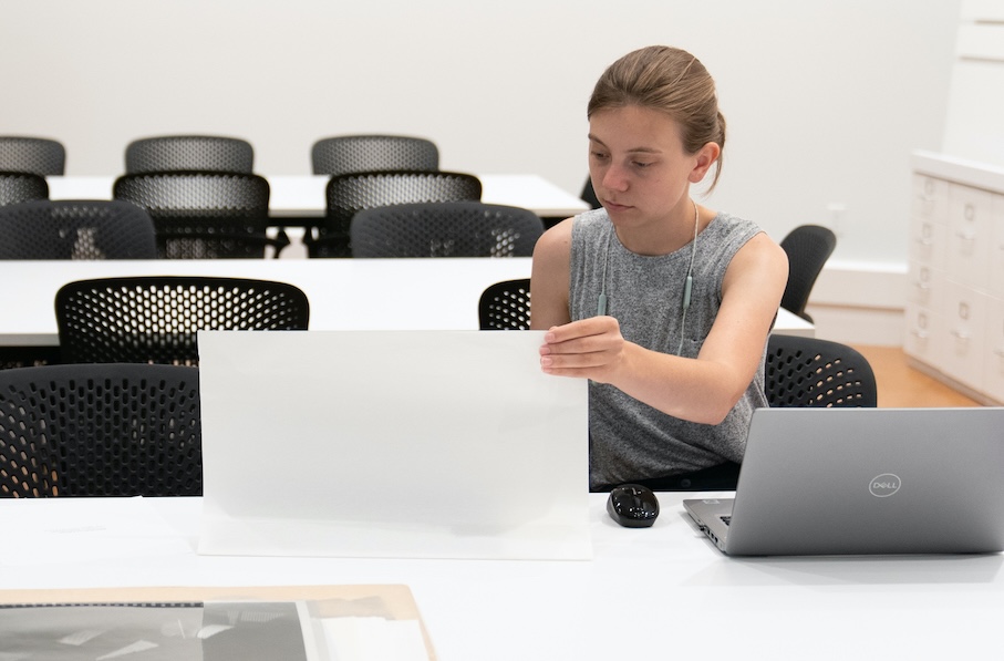 A person with dark hair works at a laptop, while also looking at a work of art that is next to them on the table.