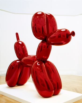 Large shiny red balloon dog sculpture sits on a white platform in a gallery. The dog reflects other artworks in the gallery on its surface in a distorted fashion. 