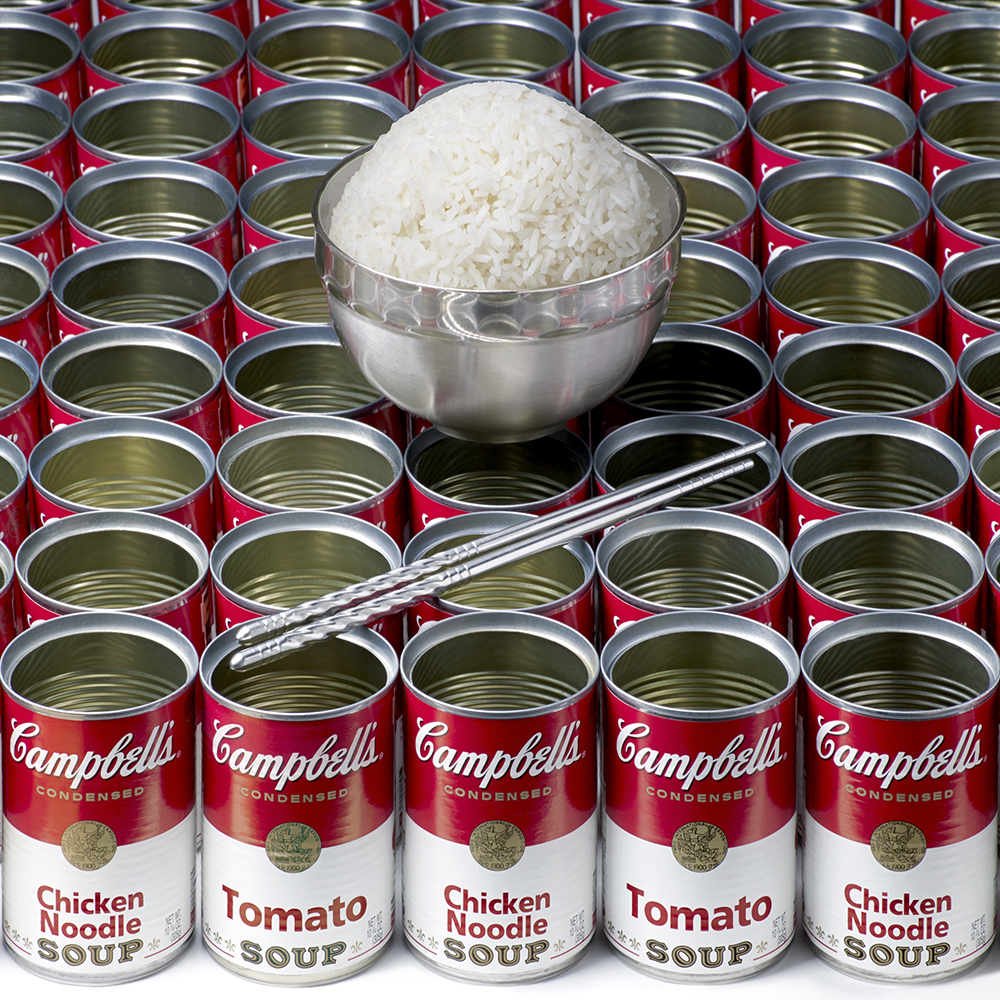 A pair of silver chopsticks and a metal bowl filled with white rice sits atop many empty cans of Campbell's Chicken Noodle Soup.