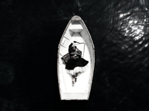 A black and white scene shows a small white rowboat adrift in the sea. A dark-skinned woman with braided hair, wearing a black dress with white petticoats, is laying on the floor of the boat, one arm raised above her head and holding the handle of a whip. Her feet are bare, and around her ankles white rope has been looped many times and tied.