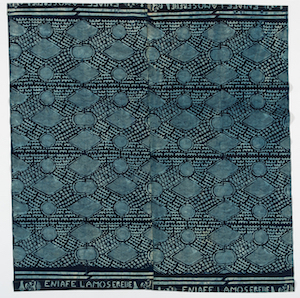 This cloth is decorated with a light blue repeated block pattern of abstract geometric forms. Along the edges is the lettering 'ENIAFE LAMOSEREIBE' and at each end of that, with axis at right angles to the lettering, there is a man's face with a hand beneath it.