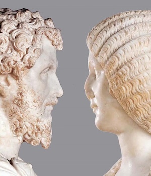 Two heads carved from light stone are facing each other. The man, on the left, has curly hair and a beard. The woman wears a tightly crimped hairstyle and has a broken nose. 