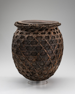 An ovoid vessel with a short neck and an outward curved rim. The clay vessel itself has patterned bands running horizontally around the body and there is an open interlaced plant fiber basket structure that surrounds the entire vessel surface. This basket structure ends at the rim with circular wrapped fiber around fiber core tightened into neck space.