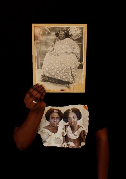 A person stands while holding in their hand a black-and-white portrait photograph that conceals their face. They wear a shirt printed with a picture of two people posed next to one another.