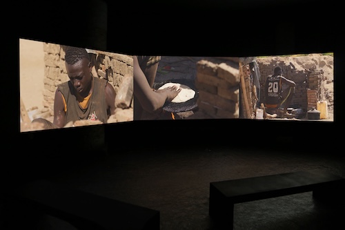 A dark room with a rectangular bench in the foreground and three connected screens behind it. The screens depict a dark-skinned man in front of a mud-brick wall, a dark-skinned arm pressing a circle of dough onto a metal cooking surface with a flame beneath it, and a dark-skinned man wearing a sports jersey crouching in a mudbrick structure surrounded by pots and plastic jugs.