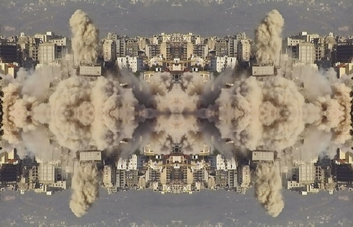 A repeated kaleidoscopic image depicts beige and white residential buildings against a blue and grey mountainous backdrop with beige plumes of smoke and dust emerging from center. The image is mirrored over the center horizon.