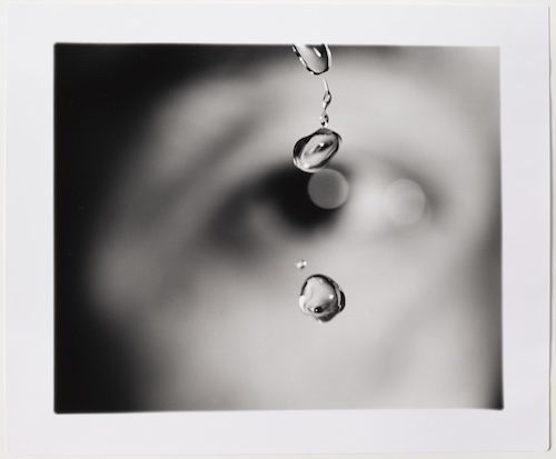 A black and white photograph focused on falling water droplets in front of an out-of-focus face, centered on a human eye.