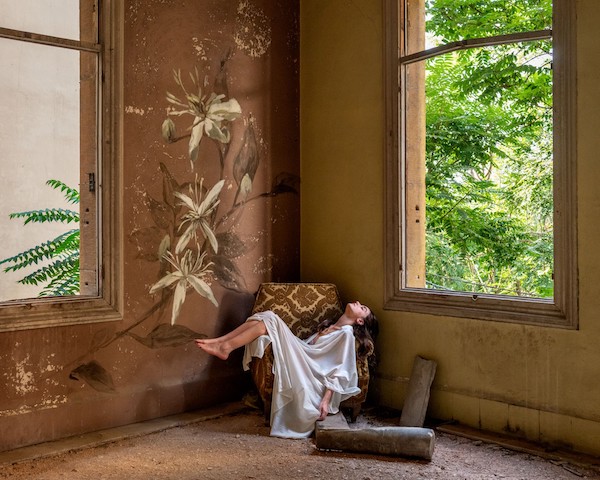 A young woman in a loose, white gown is draped across a brown upholstered chair in the corner of a decrepit room, her legs arched over the left armrest and her hair cascading over the right. Tall, open windows on either side reveal green foliage as dirt and detritus are scattered on the floor.