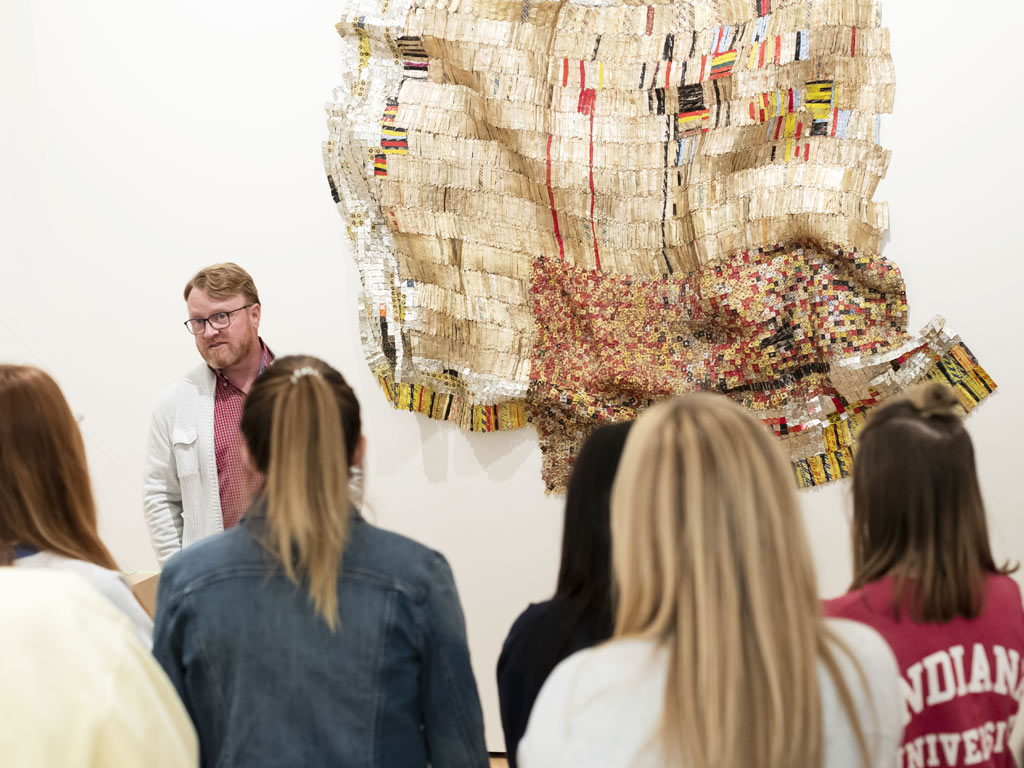 A man stands before a large multi-colored sculpture hanging on a white wall.  He is speaking to a group of students.