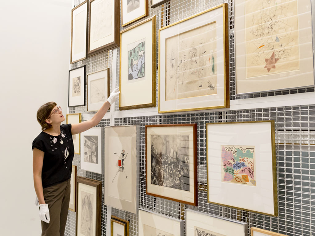 Many prints and drawings in frames hang on a large wire storage rack.  A young woman is looking at and pointing to them.