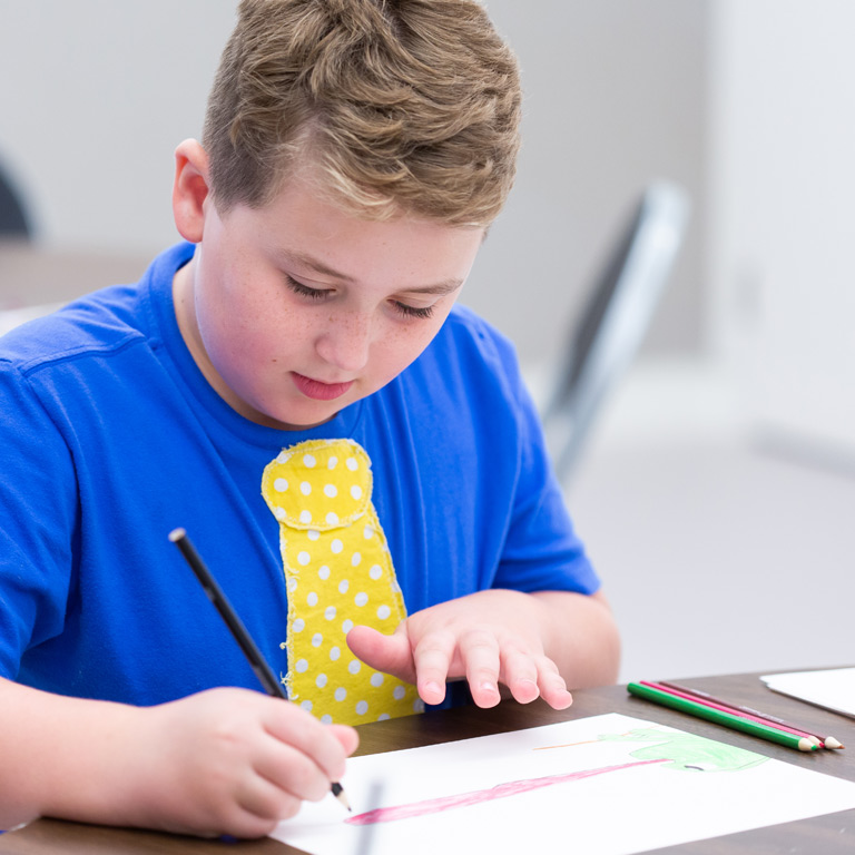 A young boy in a blue shirt is drawing at a table.