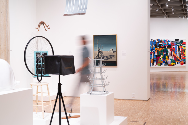 An installation view of a museum gallery shows a group of modern sculptures on white pedestals, including a bicycle wheel affixed to a wooden stool, a hat rack suspended from the ceiling, and a covered object on a black tripod. An out-of-focus person walks through the gallery past the art.
