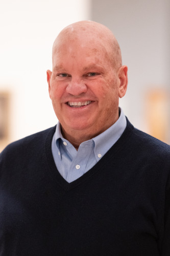 A person smiles towards the viewer in this portrait. They are visible from the shoulders up. They are fair skinned, have a shaved head, and wear a dark blue sweater with a collared blue shirt beneath. 